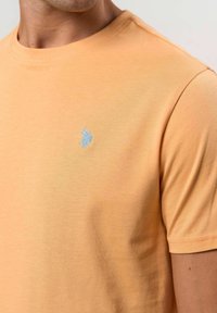 Férfi rövid ujjú világos narancssárga pólót visel, bal mellen egy kicsi, világoskék hímzett logóval.