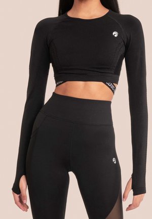 Frau trägt schwarzen langärmeligen Crop-Top und hoch taillierte schwarze Leggings mit Daumenlöchern und kleinem weißem Logo auf Brust und Oberschenkel.