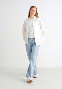 Chemise blanche à manches longues avec boutons superposée sur un t-shirt blanc, associée à un jean bleu clair et des baskets blanches. Design simple et minimaliste.