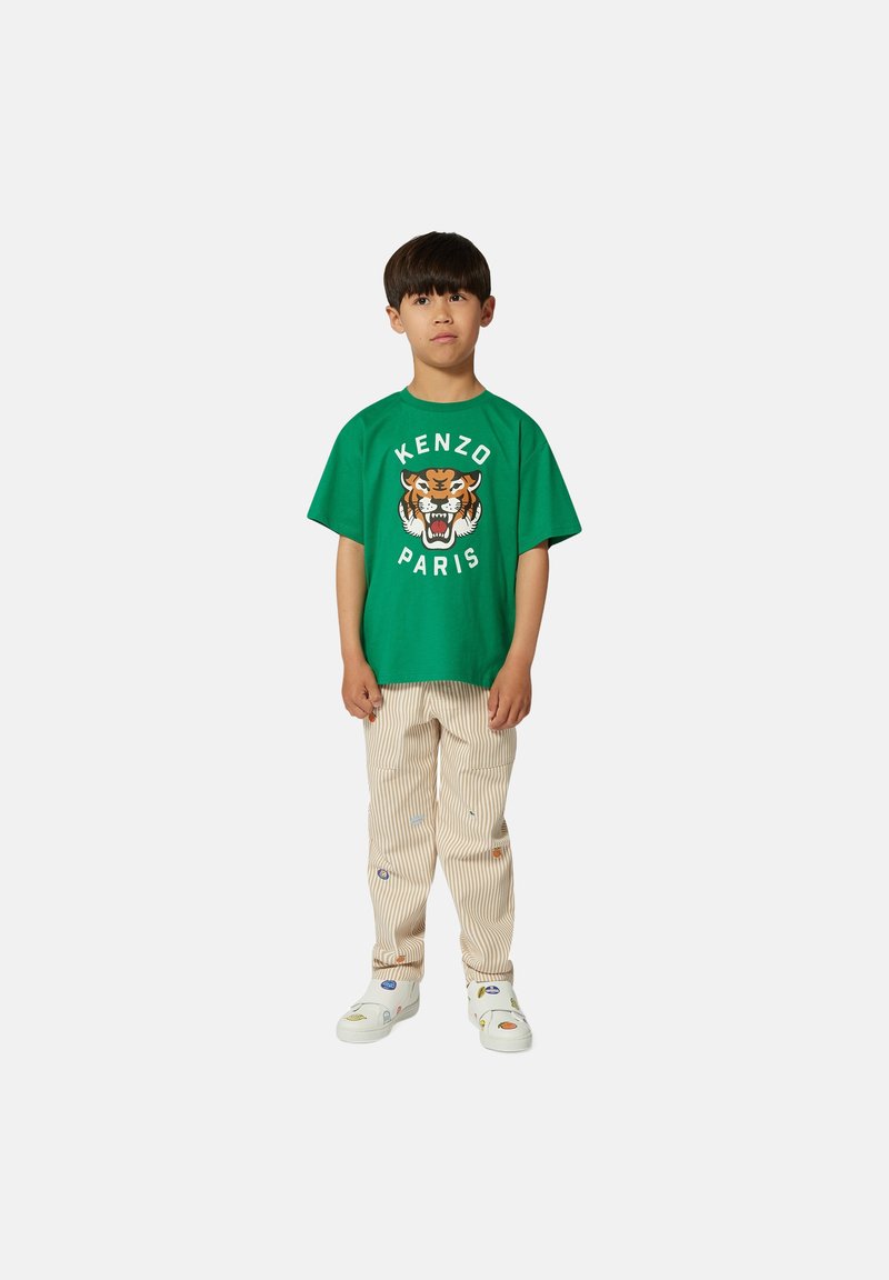 T-shirt en coton vert avec un motif de tigre et le texte "KENZO PARIS", associé à un pantalon rayé clair et des baskets blanches avec des accents colorés.
