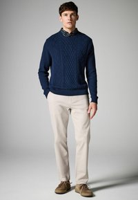 Pulover tricotat în nuanță bleumarin, cu modele texturate în formă de diamant, asortat cu pantaloni bej deschis și pantofi din piele sueded maro. Look casual, cu straturi.