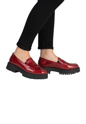 Personne portant des mocassins rouges brillants avec des semelles épaisses noires et un pantalon noir ajusté, debout sur un fond blanc.