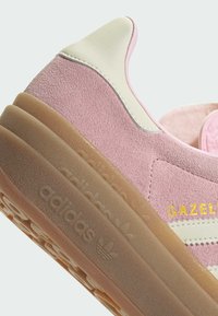 Ružové semišové tenisky s bielym koženým akcentom, textúrovanou gumovou podrážkou a zlatým brandingom 'GAZEL' pri pätke. Vyznačujú sa zaoblenou špičkou.