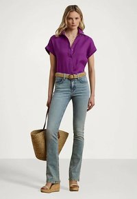 Kvinne iført en lilla kortermet bluse, lyseblå jeans med et flettet belte, brune kilehæler, og bærer en stor flettet totebag.