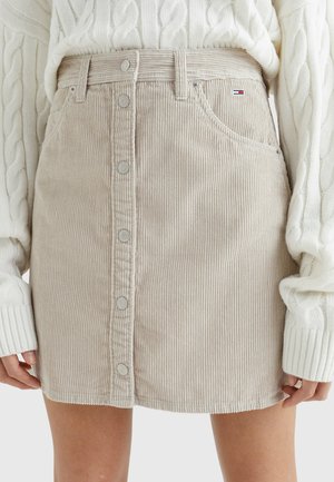 Beige cordfløjls-mini nederdel med knapper foran, båret sammen med en hvid kabelstrikket sweater, der viser forside detaljer og et lille mærkelogo på lommen.