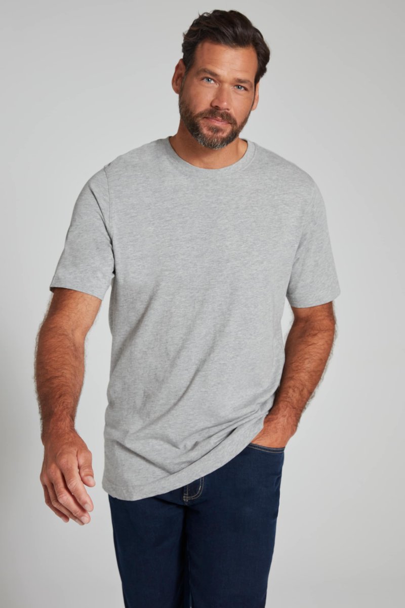 Hombre con barba y cabello oscuro, vestido con una camiseta de manga corta gris clara y vaqueros azul oscuro, posando frente a un fondo liso y claro.
