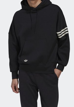 Schwarzer Hoodie mit Kängurutasche, drei weißen Streifen am linken Ärmel und kleinem weißen Adidas-Logo nahe der Tasche, getragen mit schwarzer Hose.