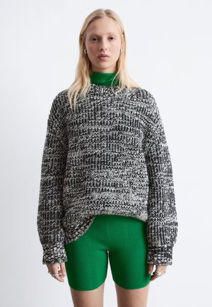 Młoda osoba o blond włosach ubrana w czarno-biały dziergany sweter na zielonym prążkowanym golfie i dopasowanych zielonych szortach.