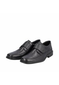 Chaussures en cuir noir avec une finition lisse, bout carré et sangle réglable. Semelle en caoutchouc robuste avec des accents de couture visibles.