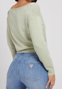 Jasnozielony sweter z szerokim dekoltem i ściągaczami na rękawach, zestawiony z niebieskimi dżinsami z naszywką w kształcie trójkąta na tylnej kieszeni.