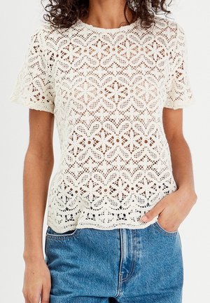 Femme portant un haut en dentelle blanche à manches courtes avec des motifs floraux et un jean en denim bleu, une main dans la poche.