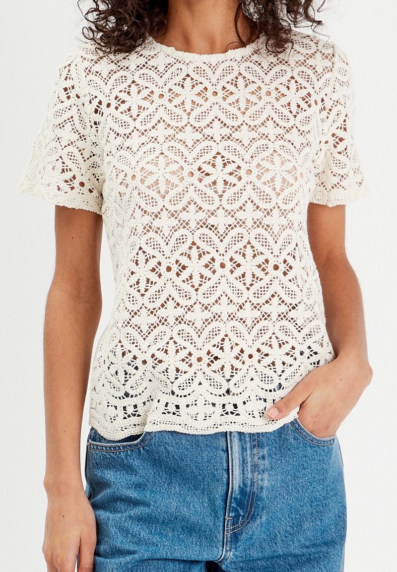 Femme portant un haut en dentelle blanche à manches courtes avec des motifs floraux et un jean en denim bleu, une main dans la poche.