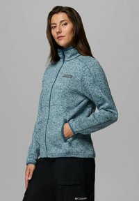 Giacca di pile blu con colletto alto, zip frontale e due tasche laterali. Presenta una texture mélange e un logo sulla parte sinistra del petto.
