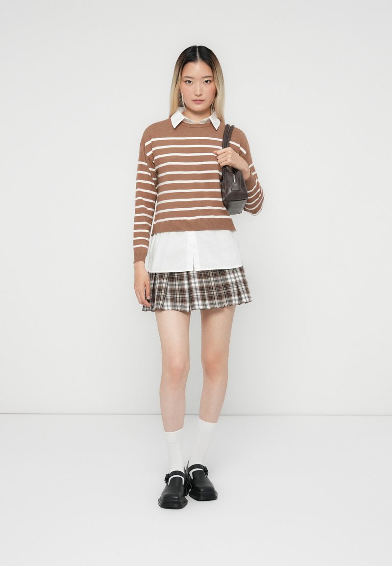 Maglione a righe marroni e bianche sopra una camicia bianca, abbinato a una mini gonna a quadri. Gli accessori includono una piccola borsa nera e scarpe nere.