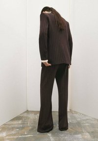 Completo marrone a righe con pantaloni svasati e un blazer lungo e oversize, mostrato di spalle su uno sfondo minimalista.