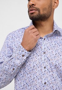 Camicia azzurro chiaro con bottoni, caratterizzata da un motivo floreale nei toni di blu e marrone, con colletto a punta e bottoni marroni.