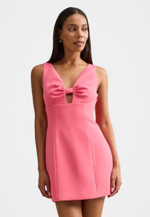 Femeie purtând o rochie mini roz fără mâneci, cu un detaliu în formă de fundă la piept, stând în fața unui fundal alb simplu.