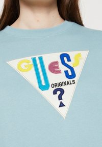 Ljust blå sweatshirt med en triangulär vit patch med färgglad text "GUESS ORIGINALS" och ett frågetecken, tillverkad av broderad tyg.