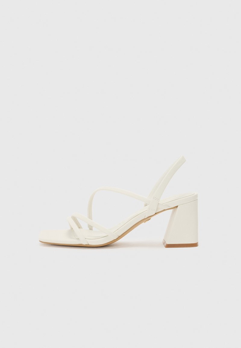 Des sandales blanches à talons présentent un bout carré, de fines lanières et un talon épais. La semelle est lisse avec une légère texture sur un fond uni.