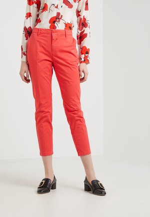 Mujer con pantalones cortos de color rojo intenso, una blusa crema con estampado floral rojo y mocasines negros de tacón con hebillas doradas.