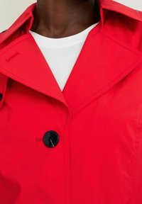 Abrigo trinchera rojo con gran cuello, botones negros y una tela suave. Presenta un detalle de logotipo en el cuello y una camiseta blanca debajo.