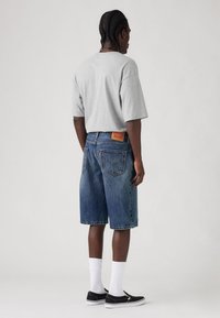 T-shirt cinzenta oversized, calções de ganga em azul claro a médio, meias brancas até ao tornozelo, sapatos pretos sem atacadores. Logo e costura notáveis no bolso traseiro.