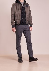 Olivgrön bomberjacka med ribbade ärmar och dragkedja, lager över en svart mönstrad hoodie och grå chinos, matchat med svarta snörskor.