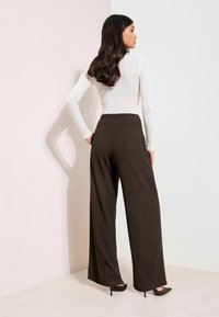 Wijde bruine broek met een geribbelde structuur, gecombineerd met een aansluitend wit topje met lange mouwen. Hoge hakken maken de look af.