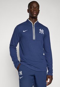 Námořnický modrý pulovr Nike s polovičním zipem, s šedým límcem, bílým logem Yankees na hrudi a dlouhými rukávy.