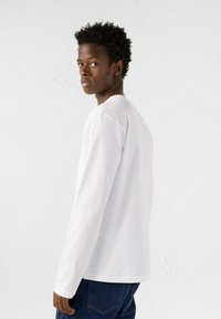 T-shirt bianco a maniche lunghe con scollo tondo, realizzato in tessuto morbido. Il modello indossa jeans blu, mostrando un design minimalista e casual.