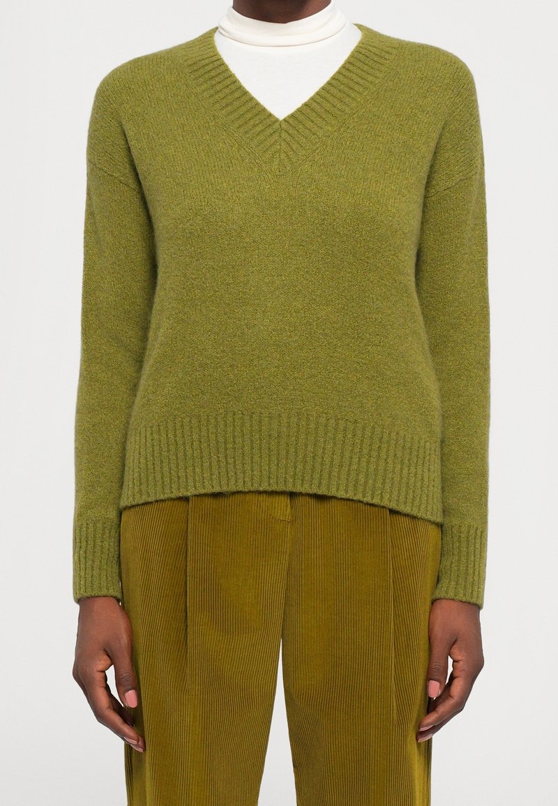 Personne portant un pull vert olive à col en V par-dessus un col roulé blanc et un pantalon en velours côtelé moutarde, debout devant un fond clair uni.