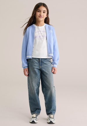 Világoskék cipzáras dzseki fehér csíkos díszítéssel, fehér pólóval, amelyen lila felirat található. Laza szabású kék farmerrel és sportcipővel viselve.