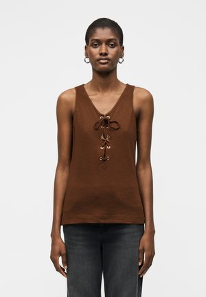 Γυναίκα με αμάνικη καφέ μπλούζα με δέσιμο μπροστά και μαύρο παντελόνι, στέκεται απέναντι σε απλό φωτεινό φόντο.