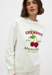 Weißes Sweatshirt mit grüner und roter Kirschen-Grafik und dem Text "Süße Kirschen 100% Natürlich Immer Frei." Rippbündchen an den Ärmeln und am Ausschnitt.