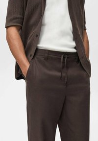 Braune leichte Hose mit einem Kordelzugbund, kombiniert mit einer strukturierten dunkelbraunen Strickjacke über einem weißen gerippten Shirt.