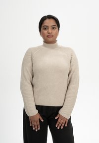 Beige Rippstrickpullover mit hohem Kragen, langen Ärmeln und strukturiertem Design. Kombiniert mit schwarzen Hosen. Weiches Material und taillierte Form.