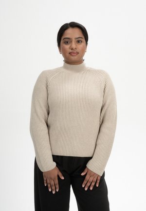 Beige geribde trui met een hoge hals, featuring lange mouwen en een textuurontwerp. Gecombineerd met zwarte pantalon. Zacht materiaal en een getailleerde vorm.