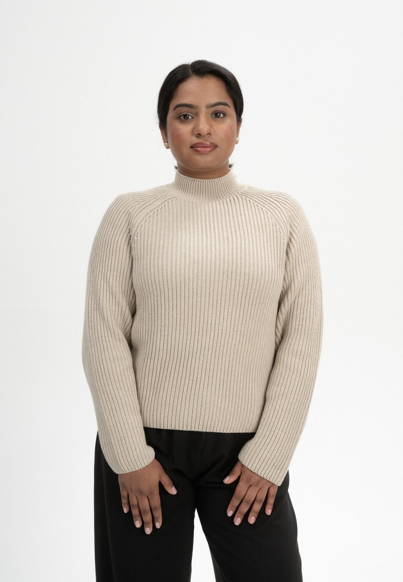 Beige Rippstrickpullover mit hohem Kragen, langen Ärmeln und strukturiertem Design. Kombiniert mit schwarzen Hosen. Weiches Material und taillierte Form.