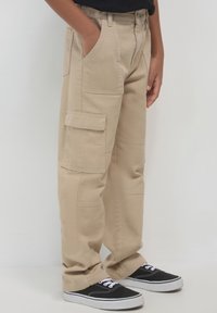 Beige Cargo-Hosen aus robustem Stoff, mit mehreren Taschen, geradem Schnitt und texturierter Oberfläche. Kombiniert mit schwarzen Sneakern.