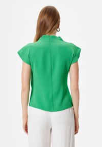 Blusa verde con maniche a cappuccio, texture liscia, dettagli delle cuciture sul retro e orlo arrotondato. Indossata con pantaloni chiari.