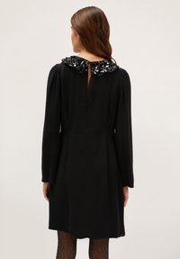 Vestido negro de manga larga con cuello decorativo de lentejuelas. Cuenta con un ojal en la espalda y cierre de cremallera. Tejido suave, diseño hasta la rodilla.
