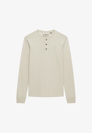 Camisa henley de manga larga en un tono beige claro, hecha de algodón, que cuenta con tres botones marrones y un pequeño logo en el lado izquierdo del pecho.