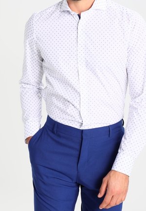 Weißes Hemd mit kleinen blauen Punkten, kombiniert mit einer schmal geschnittenen blauen Hose. Das Hemd hat einen klassischen Kragen und ein tailliertes Design.