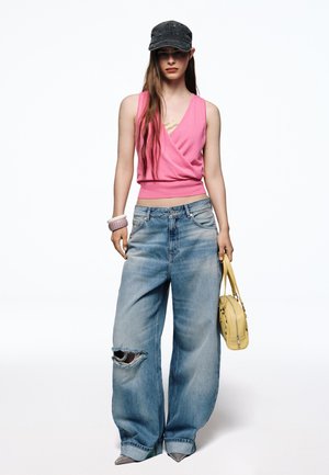 Femme portant une casquette noire, un haut rose sans manches, un jean large déchiré, tenant un sac à main jaune et portant des chaussures pointues et des bracelets.