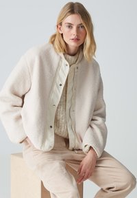 Veste en crème duveteuse avec un panneau avant en ivoire mat et fermeture à pression, portée sur un pull en tricot beige et un pantalon en velours côtelé beige clair.