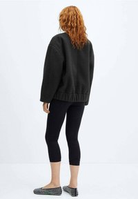 Sort fleecetrøje i oversized model med sænkede skuldre, parret med sorte leggings. Fodtøjet har et nitte-design. Baggrunden er helt hvid.