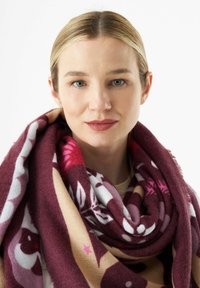 Bordeauxrode en beige sjaal met bloemenpatronen en franjes aan de randen, om de nek gewikkeld. Zachte textuur en gedurfde kleurcontrasten zijn duidelijk zichtbaar.