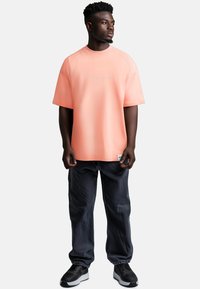 Maglietta rosa oversize a maniche corte con testo ricamato, abbinata a jeans larghi blu scuro e scarpe sportive nere.