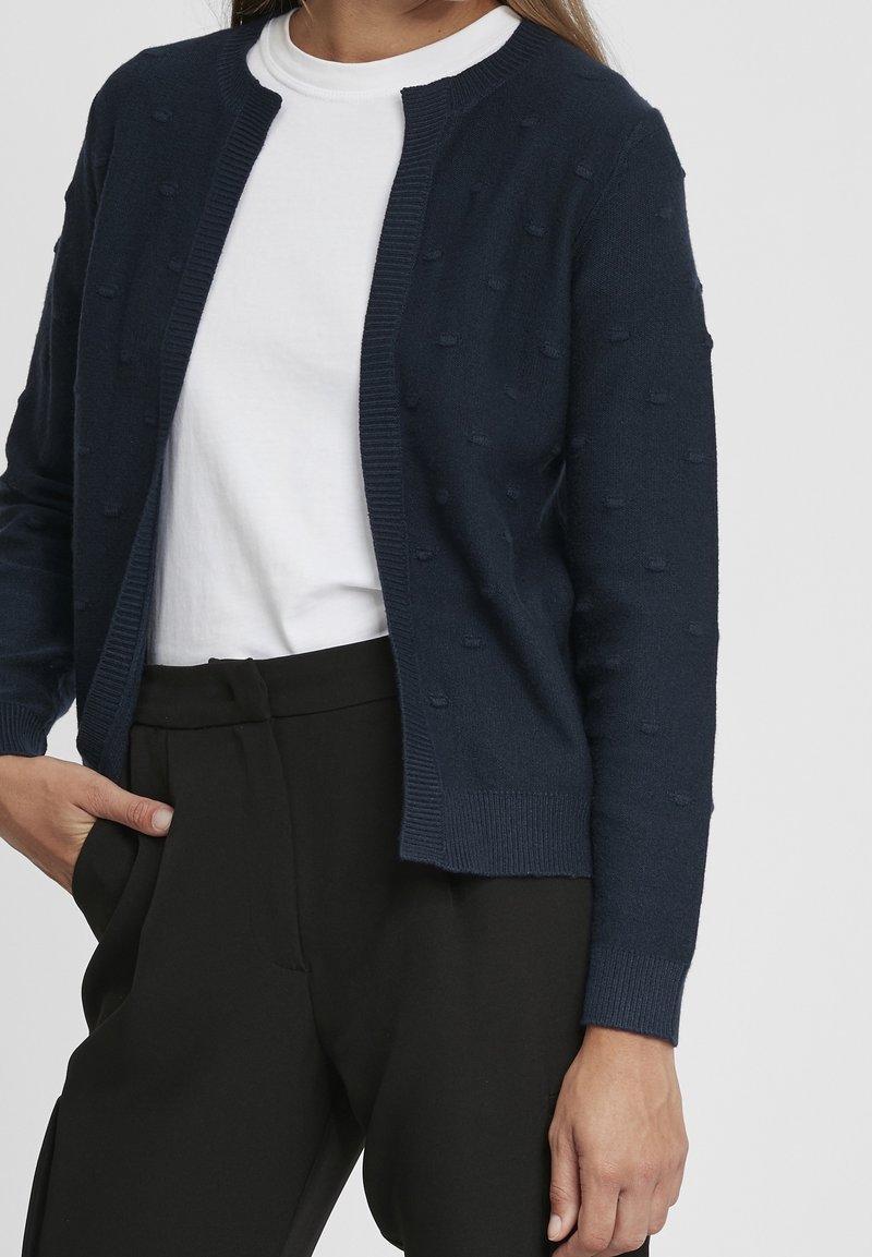 Femme portant un cardigan texturé bleu marine sur un t-shirt blanc avec un pantalon noir taille haute, posant devant un fond uni.