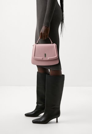 Femme en robe grise côtelée tenant un sac à main rose, portant des bottes noires montantes à bout pointu avec des talons fins, debout devant un fond blanc.